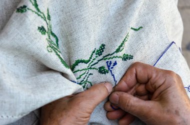 Yaşlı bir kadının elleri keten kumaşa çapraz dikişli çiçek deseni işliyor. Nakış, el işi, iğne konsepti. Yakın çekim.