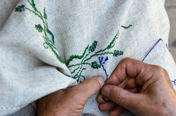 Yaşlı bir kadının elleri keten kumaşa çapraz dikişli çiçek deseni işliyor. Nakış, el işi, iğne konsepti. Yakın çekim.