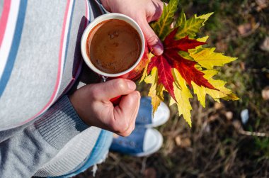Sıradan kıyafetler giymiş, elinde kahve fincanı ve renkli sonbahar yaprakları tutan bir kadın. Sonbahar havası. Hafta sonu, sonbahar tatili, eğlence konsepti. Yukarıdan bak. Yakın plan.