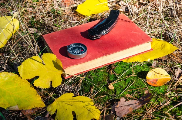 Il concetto di sopravvivenza e orientamento nella foresta. Bussola, coltello chiudibile, libro rosso sul fogliame autunnale e terreno muschiato. Di giorno, nelle giornate di sole. Primo piano — Foto Stock