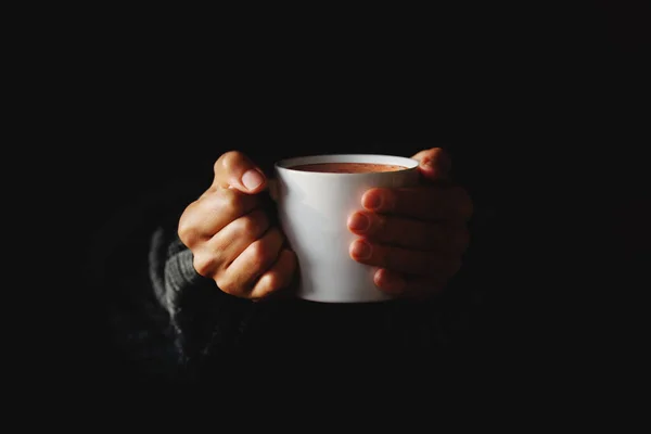 Cup of coffee in women\'s hands on black background. Woman warming her hands on mug of hot beverage in the dark. Copy space. Minimal style. Low key