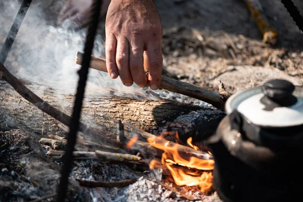 Cooking on touristic campfire. Smoked kettle and male hand throwing firewood into the fire. Touristic life concept. Selective focus