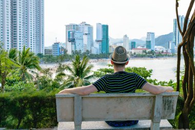 Şapkalı adam bankta oturmuş gökdelenlerle sahile bakıyor.
