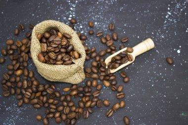 Koyu bir arka planda çuval bezi içinde kavrulmuş kahve çekirdekleri. Çantanın yanındaki tahta kürek.