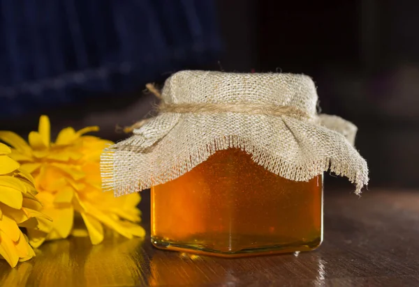 Bee honey in a glass jar with cover of burlap and yellow flowers on a dark background. Healthy food, farm products. — Stock Photo, Image