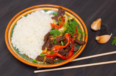 Biberli biftek ve beyaz pirinç. Renkli çizgili bir tabak, sarımsaklı karanfil, koyu arka plan ve Çin ahşap çubukları..