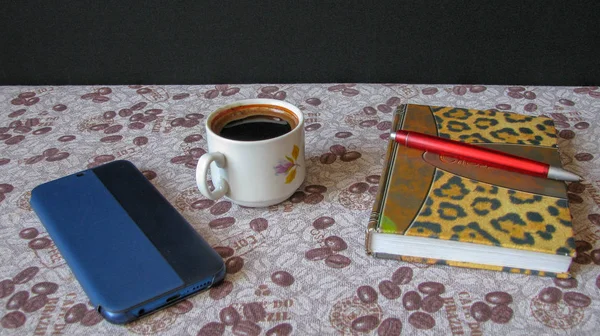 Bir fincan kahve, bir cep telefonu, bir dizüstü bilgisayar ve kahve çekirdekli bir masa örtüsü üzerinde kalem.