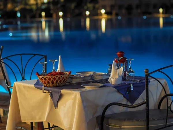 served tables at a restaurant with no people on the street preparing to welcome visitors against a backlit evening pool