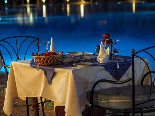 served tables at a restaurant with no people on the street preparing to welcome visitors against a backlit evening pool