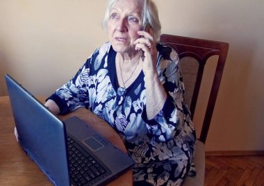 Bir dizüstü bilgisayarda çalışan ve cep telefonunda konuşan yaşlı bir kadın.
