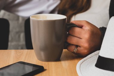 El bir fincan çay tutar. Bir arka plan görünür bir erkek eli ve bir şekil bir parçası. Bir tabloda bir kupa daha önce telefon ve bir şapka yatıyor. Fotoğraf renkli.