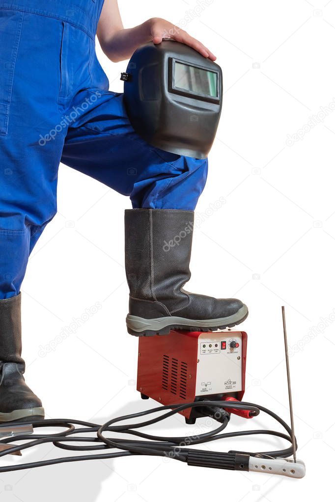The advertizing image of a lower body of the worker with the welding tool. The image of the lower part of a human body in working clothes and boots. The hand holds a welding mask. The leg is installed on the welding machine, at the very bottom lie we