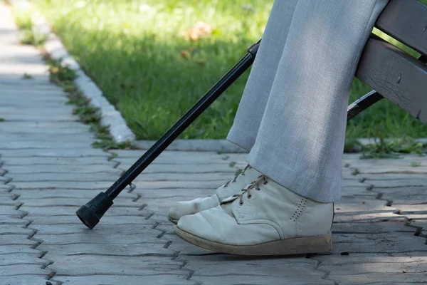 On the image of a foot, a crutch and a bench in the park. On the image of a leg in old pants, shod in old shoes, a crutch and a bench in the park.