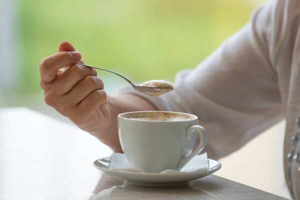 A cup of cappuccino coffee with a hand holding a spoon with foam over it.