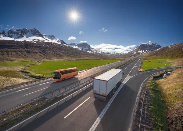 Modern bus and semi truck with container on highway driving through the mountain range towards the sun. Transportation concept.
