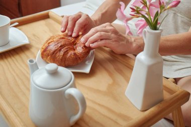 Kadın yatakta kahvaltı yapıyor, bir parça kruvasan alıyor.