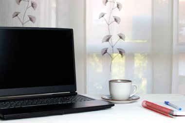 Siyah ekranlı dizüstü bilgisayar beyaz tahta masa, kahve fincanı, defter. Düzenli / Rahat iş yeri konsepti.