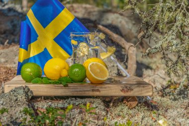 Yaz içecekleri. İskandinav doğası ve İsveç bayrağının arka planında bir bardak su, limon, nane ve nane. Farklı su damlaları kombinasyonları. İsveç