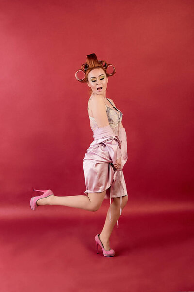 Young redhead woman with hair curlers dressed in neglige, peignoir, dressing gown posing on a burgundy background.
