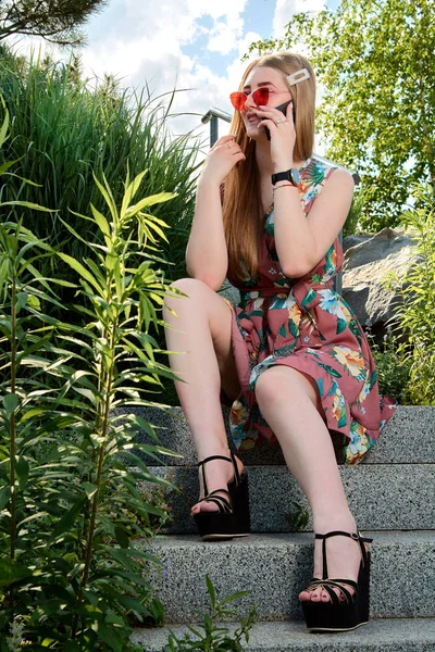 Νέα ελκυστική γυναίκα. Κόκκινα γυαλιά ηλίου, έγχρωμο φόρεμα, κινητό τηλέφωνο, 5g, 4G, 3G, 2G, δίκτυο LTE. — Φωτογραφία Αρχείου