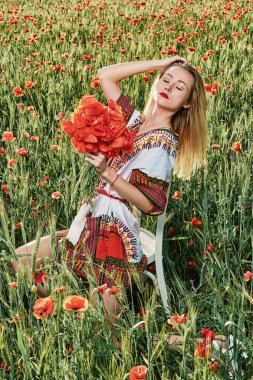 Yeşil buğday ve yabani haşhaş bir alanda beyaz kısa elbiseli uzun saçlı sarışın genç kadın.