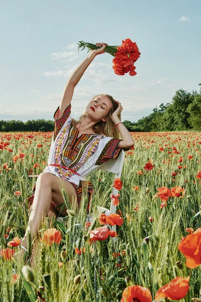 Yeşil buğday ve yabani haşhaş bir alanda beyaz kısa elbiseli uzun saçlı sarışın genç kadın.