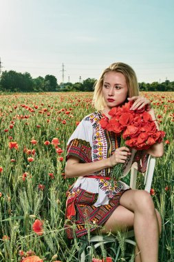 Yeşil buğday ve yabani haşhaş bir alanda beyaz kısa elbiseli uzun saçlı sarışın genç kadın.