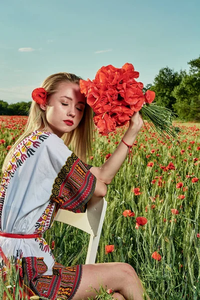 Yeşil buğday ve yabani haşhaş bir alanda beyaz kısa elbiseli uzun saçlı sarışın genç kadın.