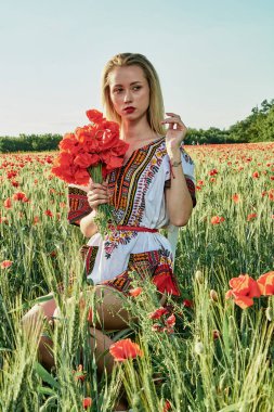 Yeşil buğday ve yabani haşhaş bir alanda beyaz kısa elbiseli uzun saçlı sarışın genç kadın.