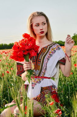 Yeşil buğday ve yabani haşhaş bir alanda beyaz kısa elbiseli uzun saçlı sarışın genç kadın.