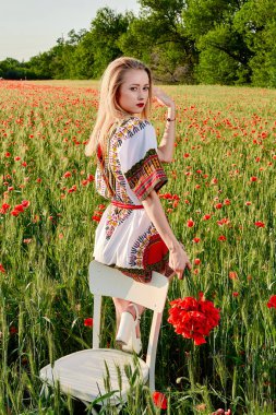 Yeşil buğday ve yabani haşhaş bir alanda beyaz kısa elbiseli uzun saçlı sarışın genç kadın.