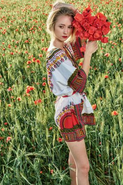 Yeşil buğday ve yabani haşhaş bir alanda beyaz kısa elbiseli uzun saçlı sarışın genç kadın.