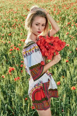 Yeşil buğday ve yabani haşhaş bir alanda beyaz kısa elbiseli uzun saçlı sarışın genç kadın.