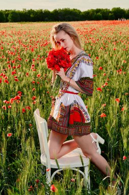 Yeşil buğday ve yabani haşhaş bir alanda beyaz kısa elbiseli uzun saçlı sarışın genç kadın.