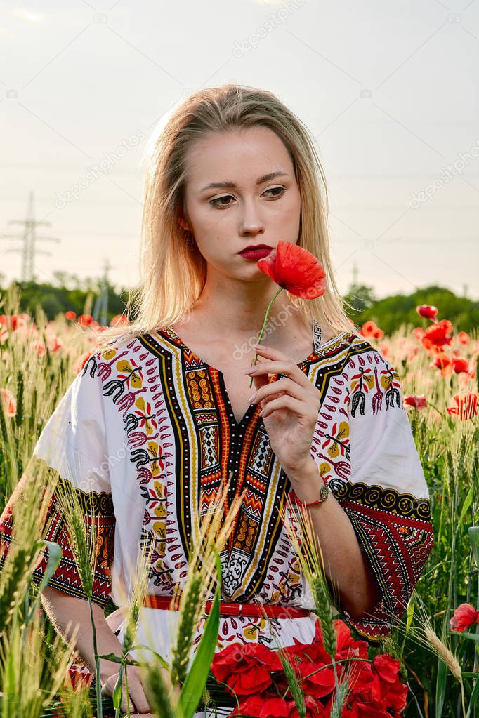 Long-haired blonde young woman in a white short dress on a field of green wheat and wild poppies.