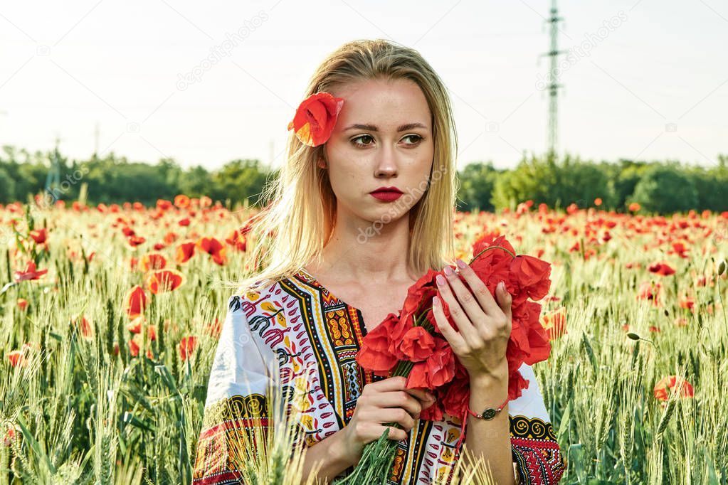 Long-haired blonde young woman in a white short dress on a field of green wheat and wild poppies.