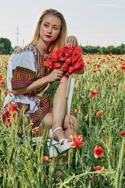 Yeşil buğday ve yabani haşhaş bir alanda beyaz kısa elbiseli uzun saçlı sarışın genç kadın.