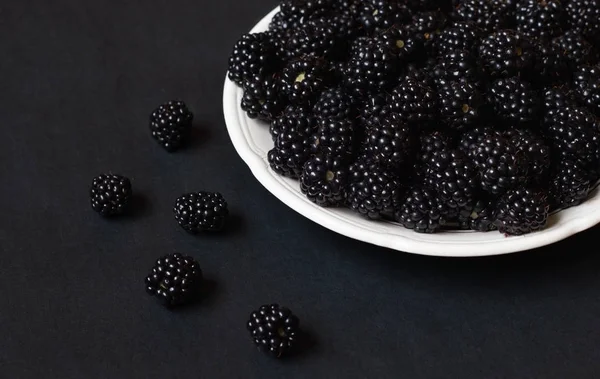 Ripe blackberries in a white plate on a black background close-up. Juicy black berries lie in darkness.