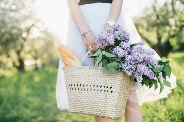 Basket with bouquet of lilacs and baguette in woman hands on bac