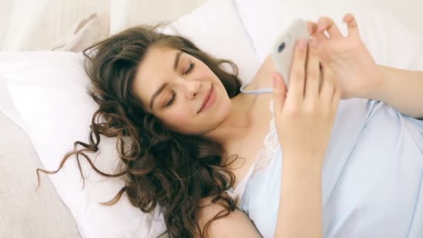 Usare il cellulare a letto. Chiacchierando umore rilassato. Donna in camera da letto soleggiata. Video filmati — Video Stock