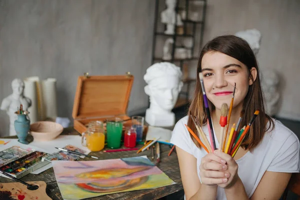 Adorable joven artista chica sosteniendo cepillos y sonriendo. Sala de taller creativo en el fondo — Foto de Stock