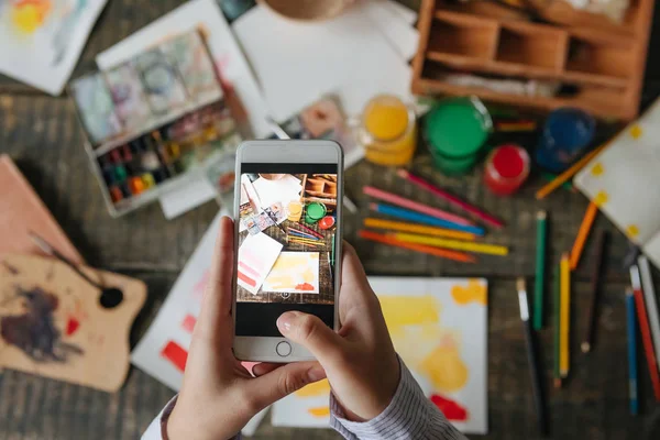 Manos de mujer sosteniendo el teléfono celular tomando fotos del trabajo de acuarela. Proceso de trabajo del artista creativo . — Foto de Stock