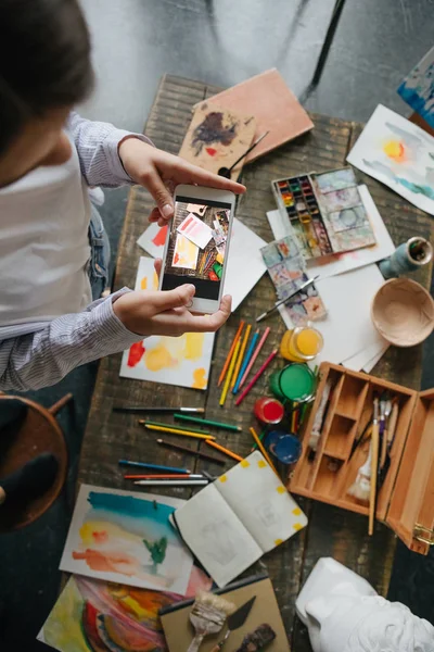 Fotografiando el proceso de dibujar imágenes en acuarela. Artista joven sosteniendo el teléfono móvil y tomando fotos . — Foto de Stock