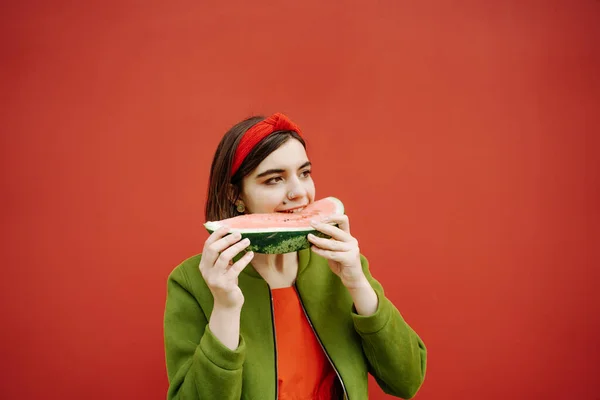 スイカを食べる若いブルネットの女の子のファッションの肖像画 ボブはヘアスタイルの赤いドレスと緑のジャケットをカット 目を閉じて食事を楽しむ 赤と緑の色表現の感情的な組み合わせ カジュアルな芸術 — ストック写真