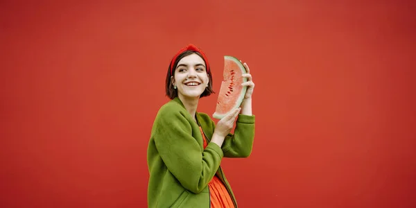 スイカと探している側のスライスを保持愛らしい幸せな笑顔の顔のブルネットの女の子 赤と緑のコントラストカラーの組み合わせ 感情的な芸術構成ジャケットとドレスの髪のファッションアクセサリー — ストック写真