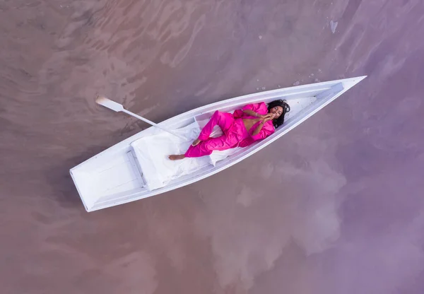 Amazing dream fairy fabulous fantasy style composition black woman in pink suit lying white boat and looking up camera. Aerial drone artistic creative conceptual imagination subconscious psychotherapy