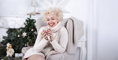 Christmas blonde woman with red lips holding cup of coffee in Christmas decorated room. looking at camera and smiling. Pretty New year eve home atmosphere. bright interior. cozy knitted winter mood
