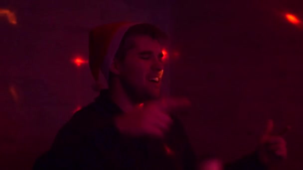 El tipo con sombrero de santa en una fiesta canta una canción. La luz parpadea como en un club. Luz multicolor. Puedes ver la botella. Año Nuevo — Vídeo de stock
