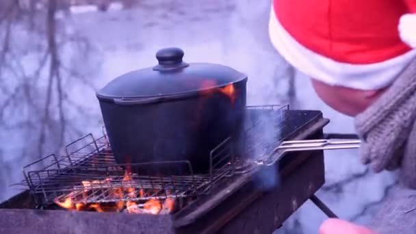 Preparation of pilaf on fire. Cooking soup. Nature, river, morning. Coldly. The guy in the hat of Santa. — Stock Video