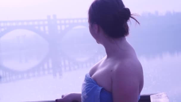 Dziewczynka w ręczniku. Rzeka, ranek, słońce, natura. Patrzy na most. Włącza się i uśmiecha się. Szczęście. — Wideo stockowe